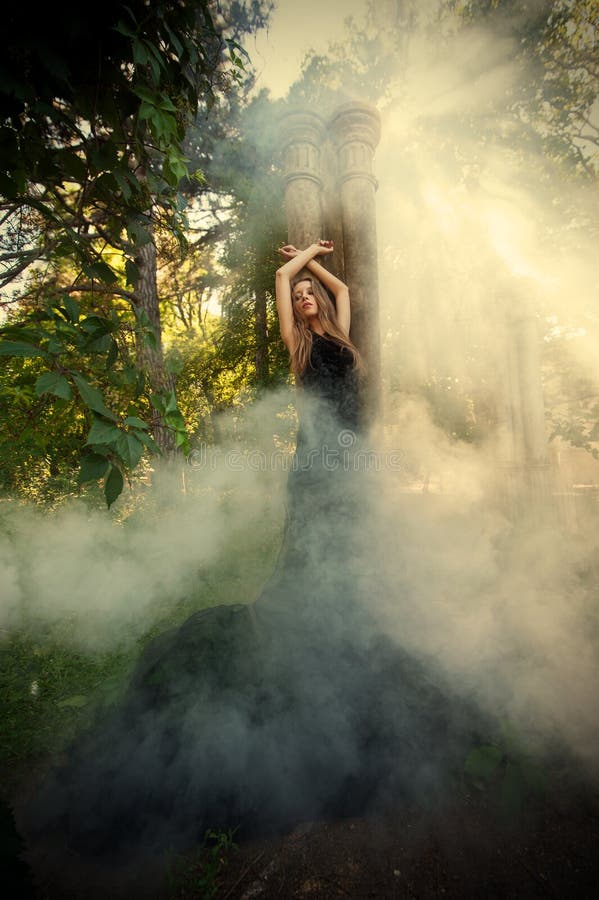 Piena lunghezza ritratto di giovane donna appoggiata alla colonna e alzando la mano nella nebbia della foresta.