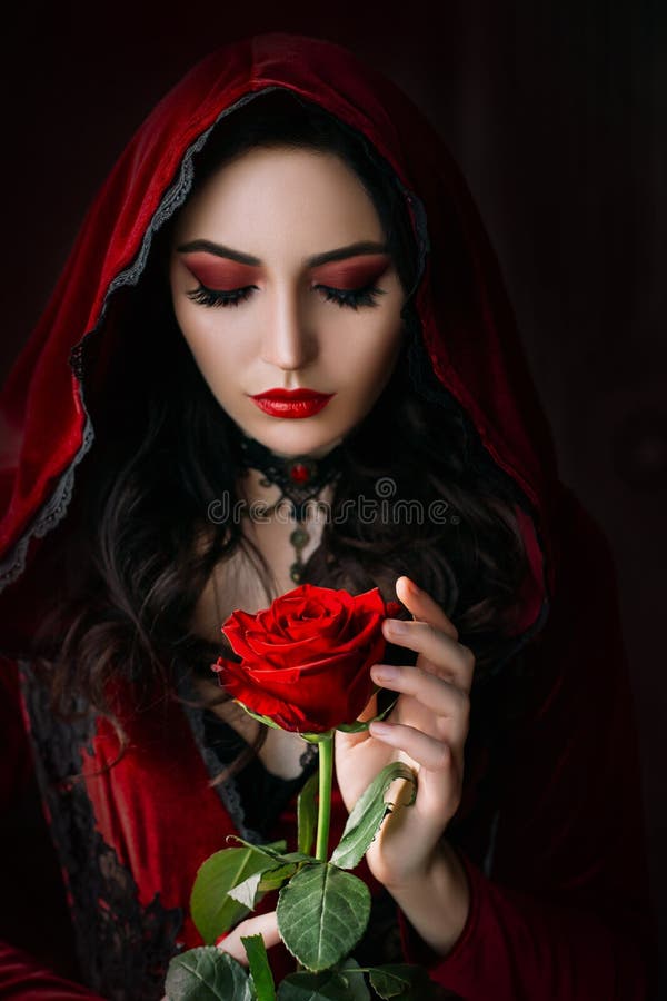 Mystical beautiful woman in a gothic costume of a medieval vampire in a hood. An adult girl holds a rose flower in her