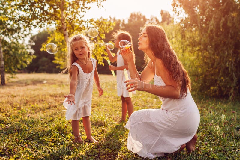 Mother helps daughters to blow bubbles in summer park. Kids having fun playing and catching bubbles outdoors. Family spending time together. Mother helps daughters to blow bubbles in summer park. Kids having fun playing and catching bubbles outdoors. Family spending time together