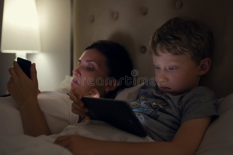 A mãe com filho olha em seus dispositivos eletrónicos que encontram-se na cama