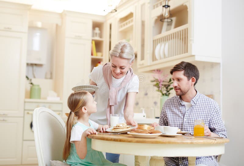 Mãe cuidadosa que põe a placa com panquecas imagem de stock Mãe cuidadosa que põe a placa com panquecas imagem de stock