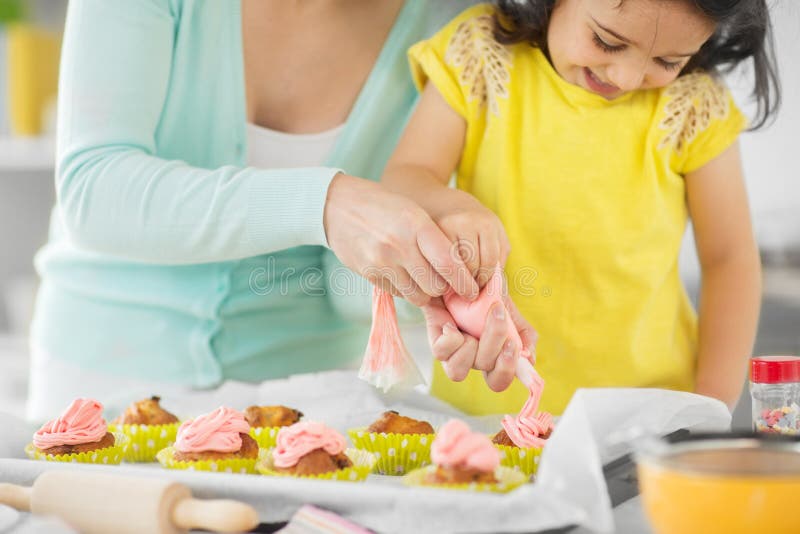 Family, cooking and people concept - mother and little daughter making and decorating cupcakes with cream frosting at home kitchen. Family, cooking and people concept - mother and little daughter making and decorating cupcakes with cream frosting at home kitchen