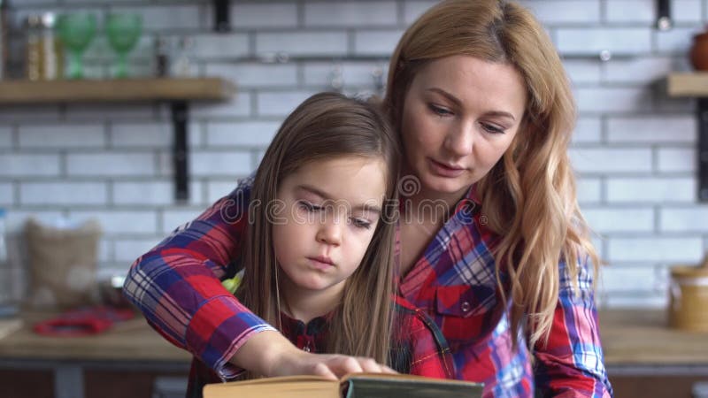 A mãe e a filha sentam-se na mesa de cozinha e na mamã que leem um livro para sua menina pequena