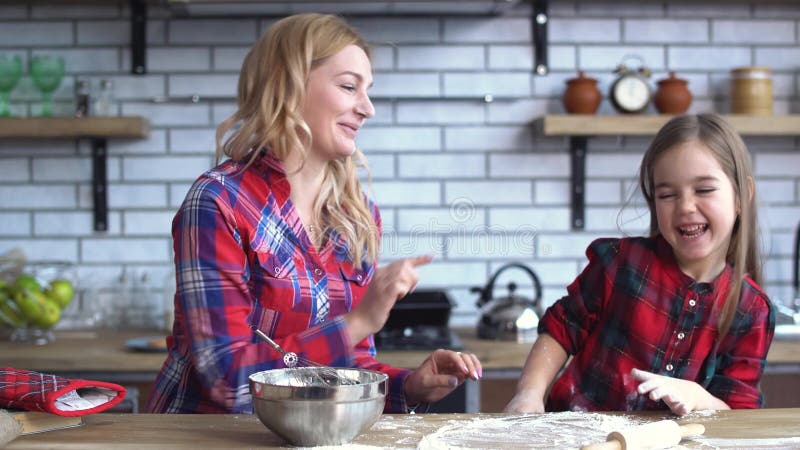 A mãe feliz com jogo da filha na cozinha, menina pequena pôs a farinha sobre a cara das mamães