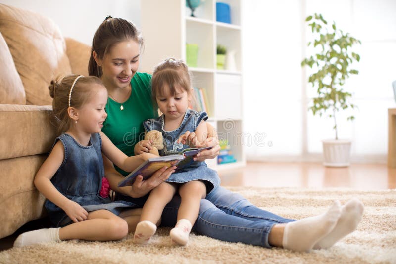 Mãe feliz e filhas pequenas que leem um livro junto na sala de visitas em casa conceito da atividade da família