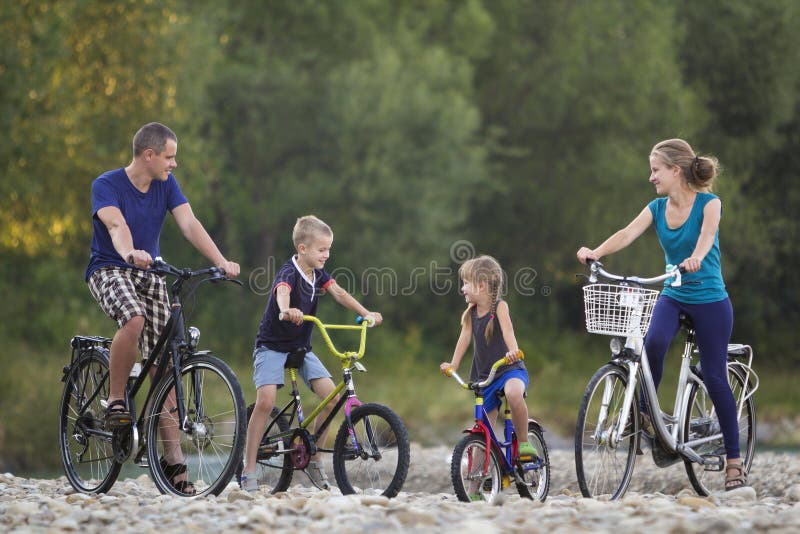 A mãe feliz nova, o pai e duas equitações louras bonitos das crianças, do menino e da menina bicycles no banco de rio pebbled no foto de stock A mãe feliz nova, o pai e duas equitações louras bonitos das crianças, do menino e da menina bicycles no banco de rio pebbled no foto de stock