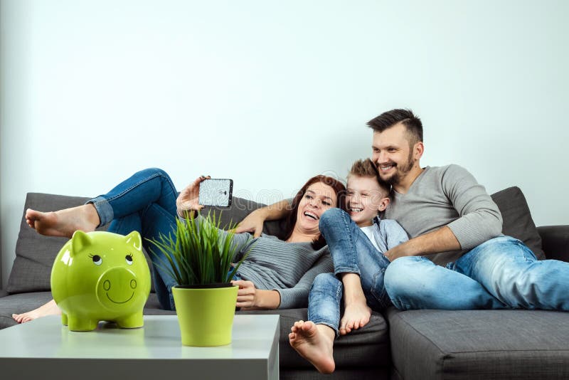 A mãe nova bonita, o pai e um filho estão fazendo o selfie usando um telefone e sorrindo ao sentar-se no sofá em casa feliz fotografia de stock A mãe nova bonita, o pai e um filho estão fazendo o selfie usando um telefone e sorrindo ao sentar-se no sofá em casa feliz fotografia de stock