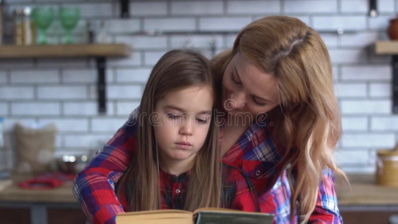 Mãe nova bonita que lê um livro para uma filha pequena bonito que senta-se na mesa de cozinha A criança é virada e