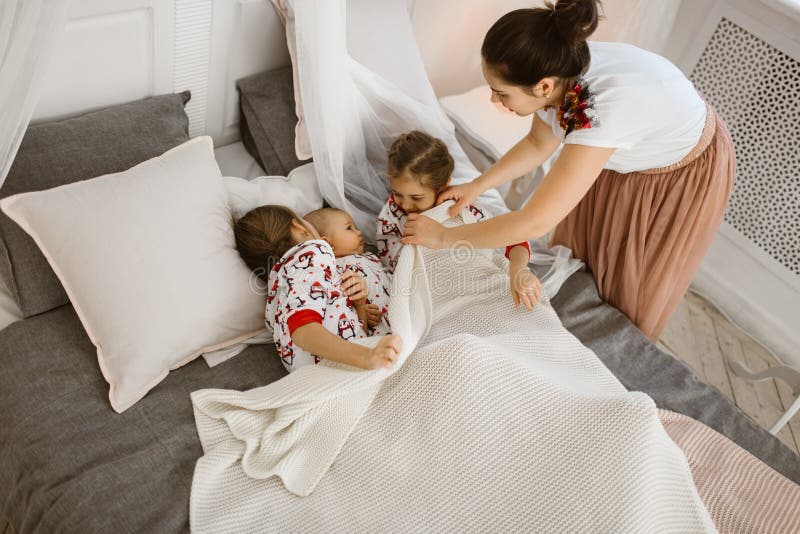A mãe nova cobrem uma cobertura branca para duas filhas e um filho minúsculo que se encontram na cama em um quarto acolhedor cl fotos de stock A mãe nova cobrem uma cobertura branca para duas filhas e um filho minúsculo que se encontram na cama em um quarto acolhedor cl fotos de stock