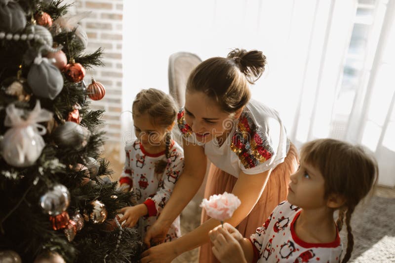 A mãe nova com as duas filhas pequenas nos pijamas decora uma árvore de ano novo na sala acolhedor com com a janela clara grand foto de stock A mãe nova com as duas filhas pequenas nos pijamas decora uma árvore de ano novo na sala acolhedor com com a janela clara grand foto de stock