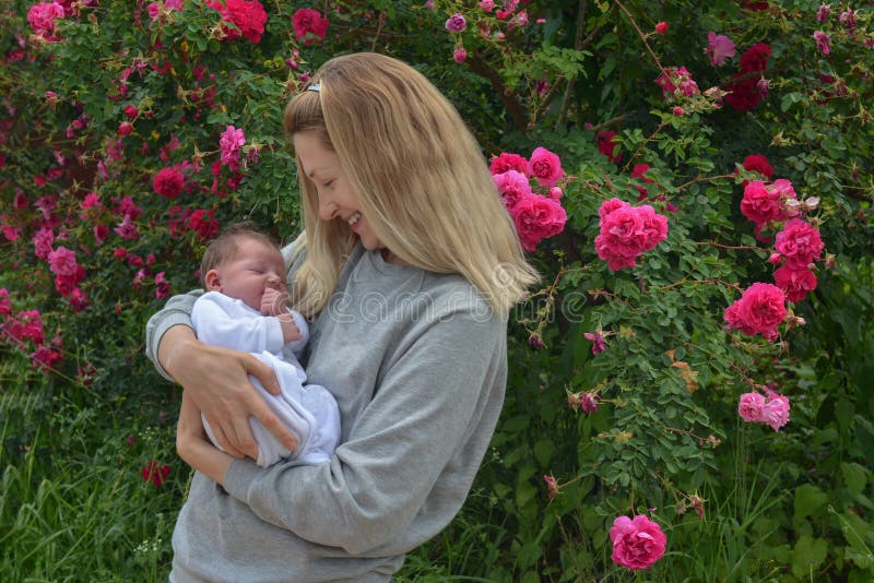 Mãe nova com uma criança recém-nascida no parque perto do arbusto com rosas imagens de stock Mãe nova com uma criança recém-nascida no parque perto do arbusto com rosas imagens de stock