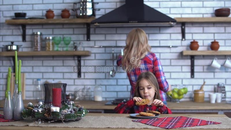 Mãe nova na camisa de manta com cabelo louro longo que cozinha o café da manhã e a cookie impertinente pequena da mordida uma da