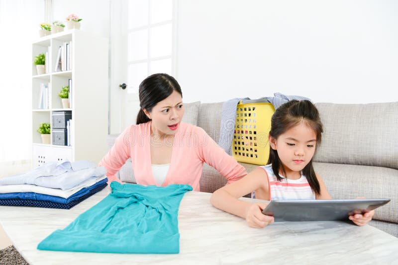 Mãe nova que encontra a menina que usa a almofada móvel. Mãe bonita nova que encontra a menina que usa o computador móvel da almofada que sente irritado quando ela que faz trabalhos domésticos na sala de visitas