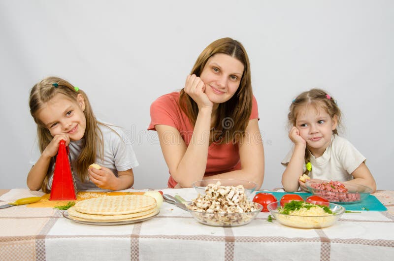 Mãe satisfeita com as duas filhas que sentam-se descansando sua cabeça em suas mãos na tabela com os produtos para a pizza fotografia de stock Mãe satisfeita com as duas filhas que sentam-se descansando sua cabeça em suas mãos na tabela com os produtos para a pizza fotografia de stock