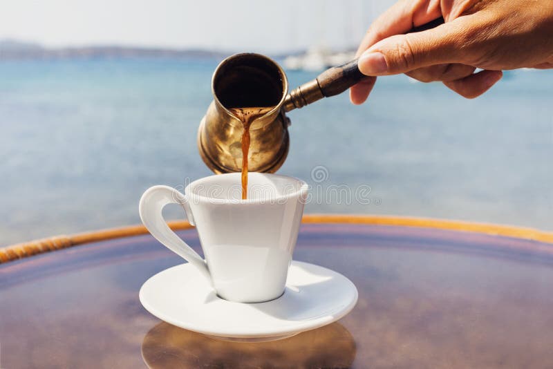 Mão fêmea que derrama o café grego tradicional em um café com um mar no fundo fotografia de stock royalty free Mão fêmea que derrama o café grego tradicional em um café com um mar no fundo fotografia de stock royalty free