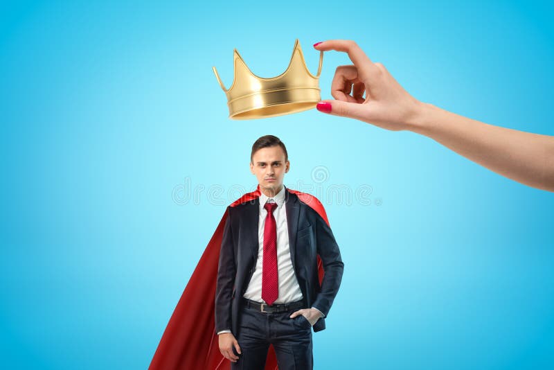 Mão fêmea que guarda a coroa dourada acima do homem de negócios novo com o casaco vermelho no fundo azul fotografia de stock royalty free Mão fêmea que guarda a coroa dourada acima do homem de negócios novo com o casaco vermelho no fundo azul fotografia de stock royalty free