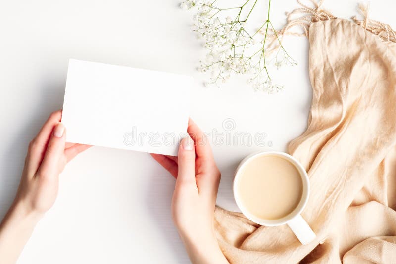 Mãos femininas segurando um cartão de papel branco sobre uma mesa branca confortável em casa com uma xícara de flores de cobertor