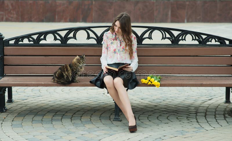 Mädchen, das ein Buch mit einer Katze auf einer Bank in der Stadt liest lizenzfreie stockbilder Mädchen, das ein Buch mit einer Katze auf einer Bank in der Stadt liest lizenzfreie stockbilder