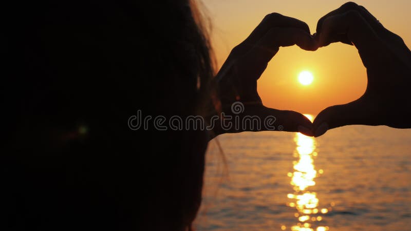 Mädchen hält Hände in Form eines Herzens auf einem Hintergrund der Sonne bei Sonnenuntergang.