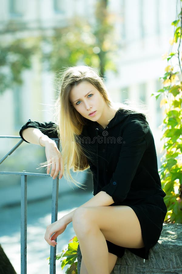 Portrait of pretty blond girl with straight long hair flowed over her shoulder posing in black casual hooded short dress sitting on stone leaning on fence on streetscape background, vertical picture. Portrait of pretty blond girl with straight long hair flowed over her shoulder posing in black casual hooded short dress sitting on stone leaning on fence on streetscape background, vertical picture