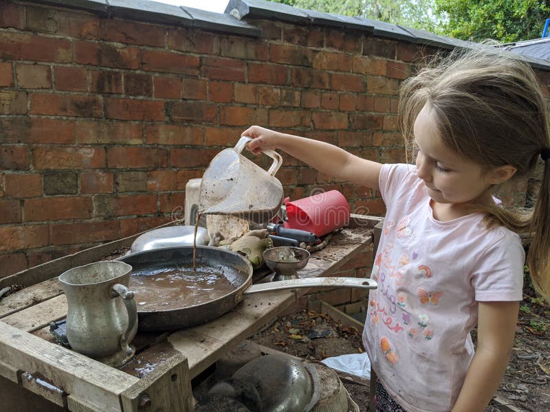 Children& x27;s sister daughter little girl long hair ponytail holding cup tipping garden red brown free outside outdoors liquid dishwater sludge pours muddy water out of plastic jug into a frying pan on a table pink t-shirt brick wall Mud kitchen recreation playing area dirty pots pans soil playground school old vintage metal wooden utensils crafted rusty discarded kids child& x27;s exploring adventure. Children& x27;s sister daughter little girl long hair ponytail holding cup tipping garden red brown free outside outdoors liquid dishwater sludge pours muddy water out of plastic jug into a frying pan on a table pink t-shirt brick wall Mud kitchen recreation playing area dirty pots pans soil playground school old vintage metal wooden utensils crafted rusty discarded kids child& x27;s exploring adventure