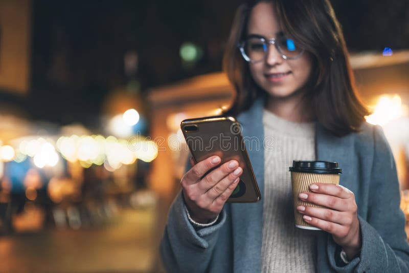 Mädchengriff im Hand-Tasse Kaffee nach draußen und mit mobilem Smartphone auf Hintergrund bokeh Licht in Nacht-Stadt Online WiFi stockfotografie