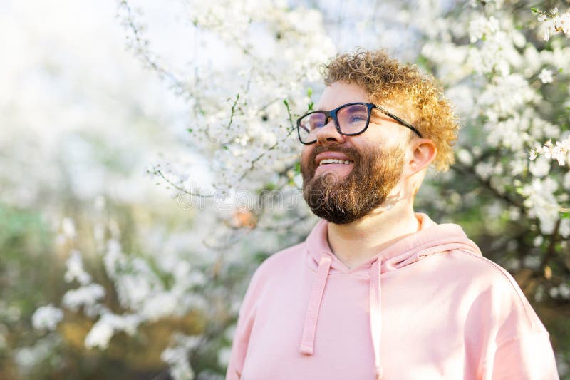 Male bearded guy standing under branches with flowers of blooming almond or cherry tree in spring garden. Spring blossom. Copy space. Male bearded guy standing under branches with flowers of blooming almond or cherry tree in spring garden. Spring blossom. Copy space