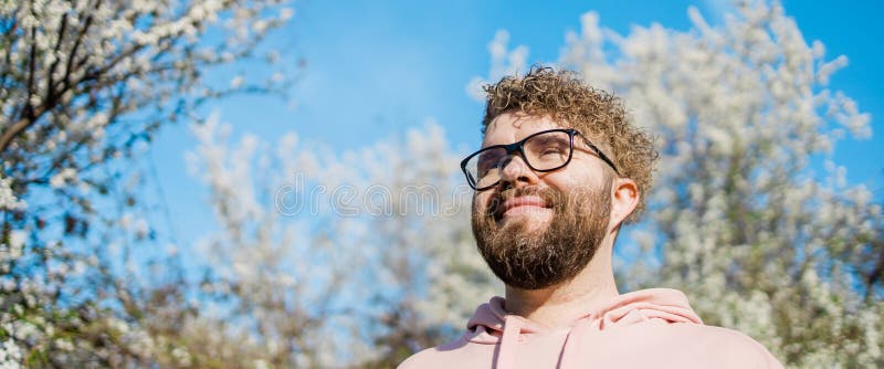 Male bearded guy standing under branches with flowers of blooming almond or cherry tree in spring garden. Spring blossom. Copy space. Male bearded guy standing under branches with flowers of blooming almond or cherry tree in spring garden. Spring blossom. Copy space