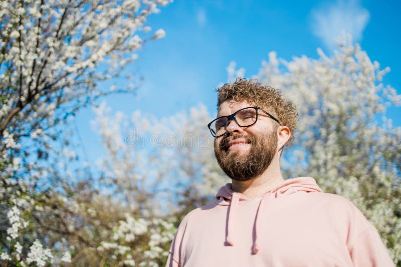 Male bearded guy standing under branches with flowers of blooming almond or cherry tree in spring garden. Spring blossom. Copy space. Male bearded guy standing under branches with flowers of blooming almond or cherry tree in spring garden. Spring blossom. Copy space