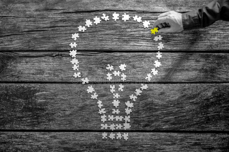 Greyscale image of male hand placing the last missing puzzle piece into a light bulb shape on a textured wooden desk with selective colour of the last puzzle, top view. Greyscale image of male hand placing the last missing puzzle piece into a light bulb shape on a textured wooden desk with selective colour of the last puzzle, top view.