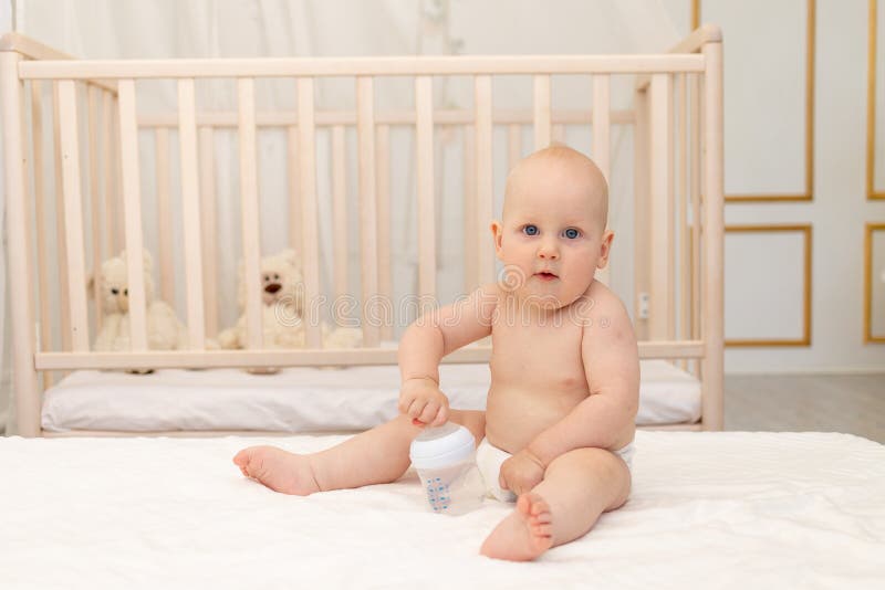 8 månaders barnpojke som sitter i blöjor på en vit bädd med en flaska mjölk på hemmabruket för barnmat första betesstället för royaltyfri bild
