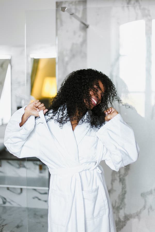 Młoda afro amerykańska kobieta z kędzierzawego włosy tanem w łazience i być ubranym białego bathrobe obraz stock Młoda afro amerykańska kobieta z kędzierzawego włosy tanem w łazience i być ubranym białego bathrobe obraz stock