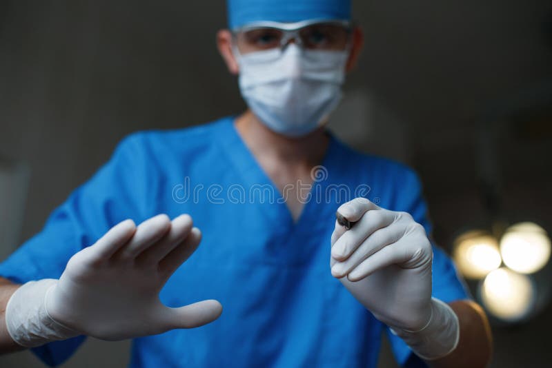 Young professional doctor in a blue medical uniform in rubber gloves in a white protective mask holds in his hand a metal scalpel in the operating room. Modern medicine and tools. Young professional doctor in a blue medical uniform in rubber gloves in a white protective mask holds in his hand a metal scalpel in the operating room. Modern medicine and tools