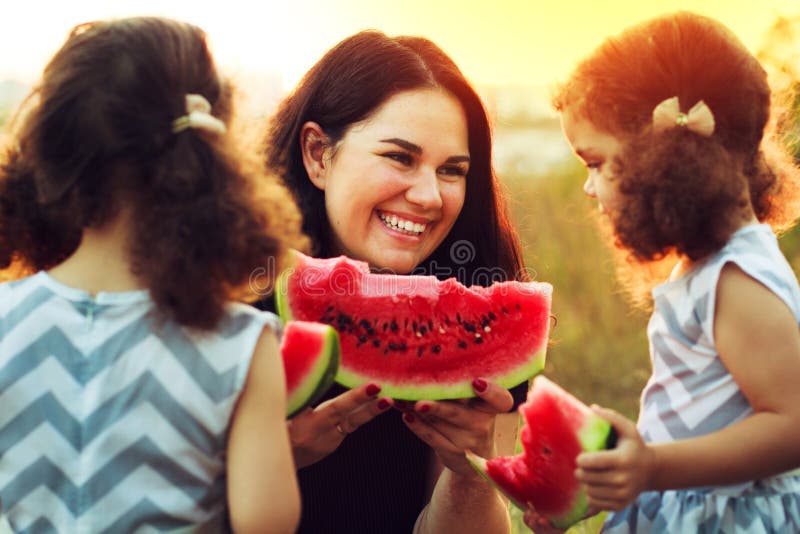 Mère attirante et petites filles jumelles mignonnes avec les cheveux bouclés appréciant la pastèque douce pendant le temps de piq photographie stock Mère attirante et petites filles jumelles mignonnes avec les cheveux bouclés appréciant la pastèque douce pendant le temps de piq photographie stock