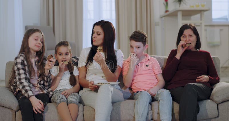 Mère attirante même avec ses enfants et mamie mangeant des biscuits tout en regardant une TV devant la caméra se reposant deda