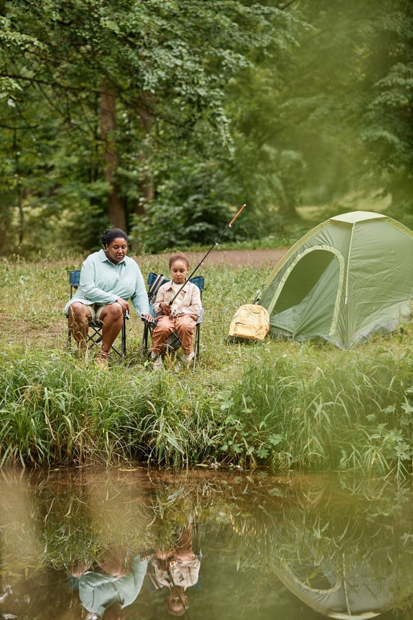 Mère et fille noire campant ensemble à l'extérieur et pêchant par rivière photo libre de droits