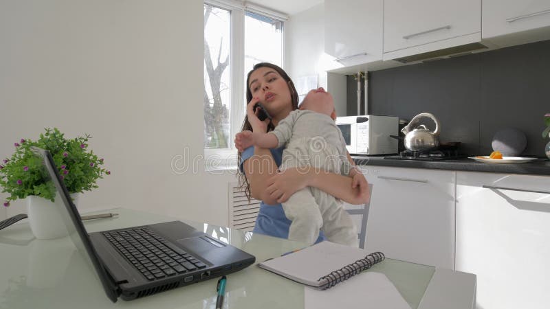 Mère multitâche avec le bébé garçon pleurant tout en travaillant sur l'ordinateur portable et parlant sur le smartphone se r