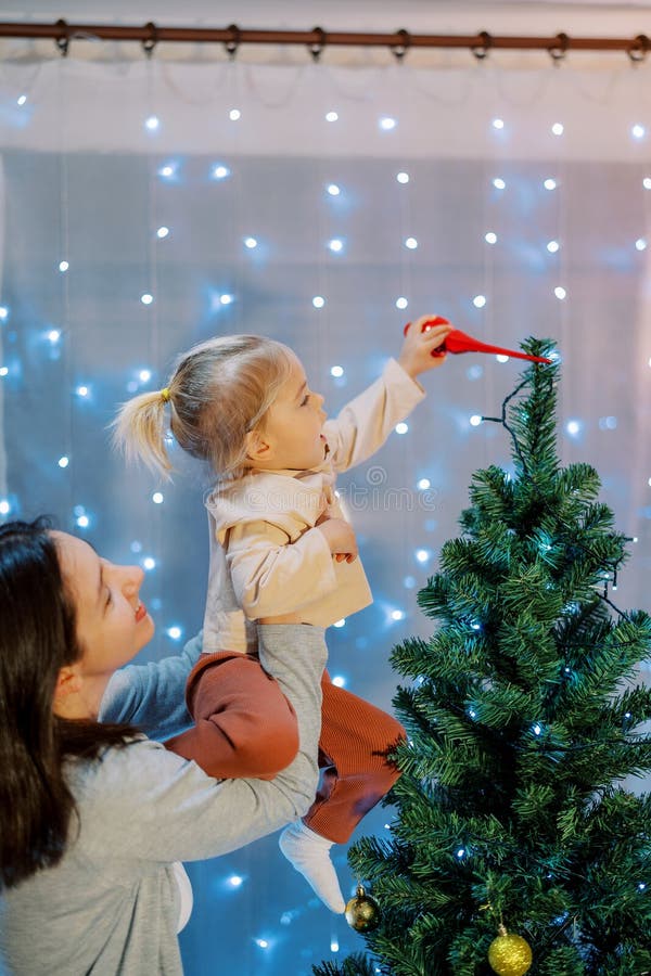 Smiling mother lifted in her arms a little girl hanging a top on a Christmas tree. High quality photo. Smiling mother lifted in her arms a little girl hanging a top on a Christmas tree. High quality photo