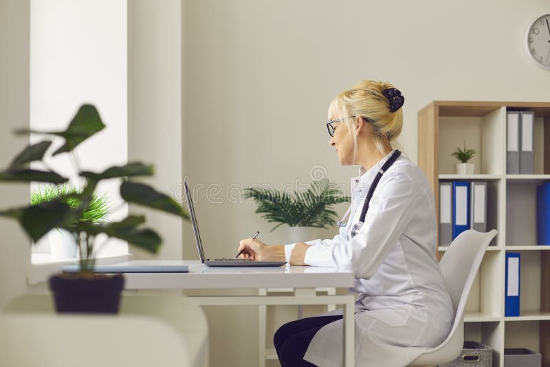 Medical routine. Side view of a busy female doctor sitting at her workplace in front of a laptop in a hospital office and writing. Doctor prescribes online prescriptions and referrals to his patients. Medical routine. Side view of a busy female doctor sitting at her workplace in front of a laptop in a hospital office and writing. Doctor prescribes online prescriptions and referrals to his patients.