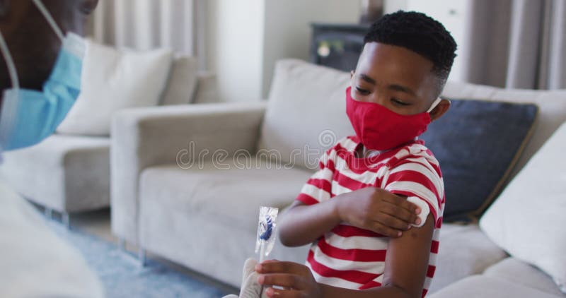 Médico da américa africana usando máscara de rosto segurando um pirulito para um menino após sua vacinação em casa