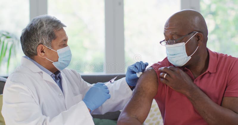 Médico masculino con máscara facial vacunando a un anciano afroamericano en casa metrajes