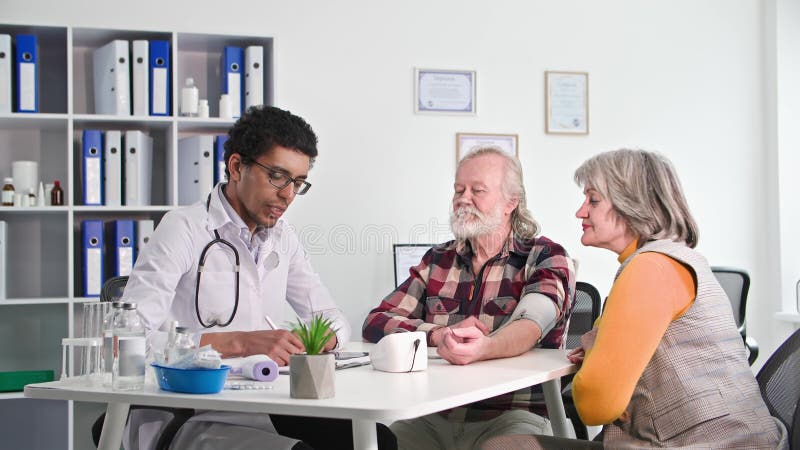 Médico negro mede a pressão de um idoso em uma recepção com sua esposa em um consultório médico
