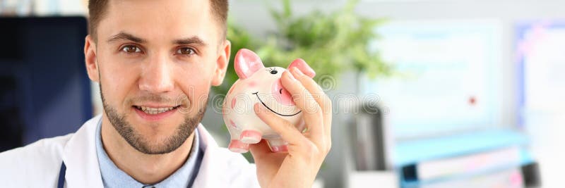Portrait of cheerful doctor shaking piggy bank. Happy physician posing in polyclinic cabinet in white uniform with stethoscope and looking at camera with certainty. Health insurance policy concept. Portrait of cheerful doctor shaking piggy bank. Happy physician posing in polyclinic cabinet in white uniform with stethoscope and looking at camera with certainty. Health insurance policy concept