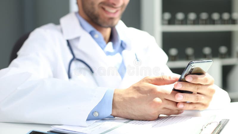 Médico varón cercano con uniforme blanco sostiene smartphone navegar por internet en la clínica. almacen de video