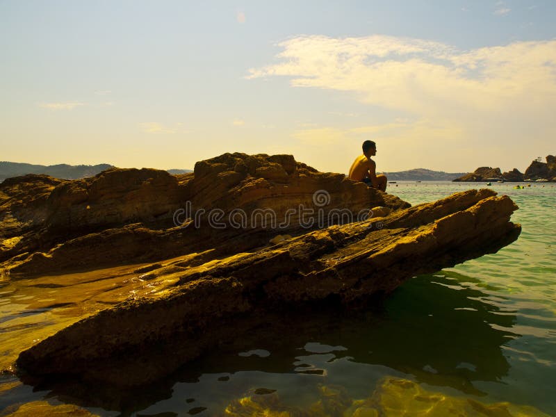 Young man sitting in a rock in the idyllic beach of Cala Fornells, Mallorca. White balance digitally modified for a creative effect. Young man sitting in a rock in the idyllic beach of Cala Fornells, Mallorca. White balance digitally modified for a creative effect