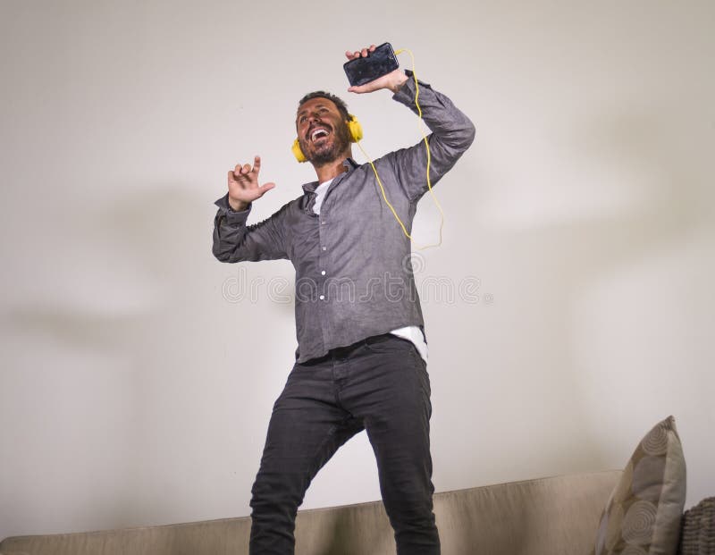 Música em linha de escuta do homem atrativo entusiasmado com fones de ouvido e música do canto do telefone celular e salto despre imagens de stock Música em linha de escuta do homem atrativo entusiasmado com fones de ouvido e música do canto do telefone celular e salto despre imagens de stock
