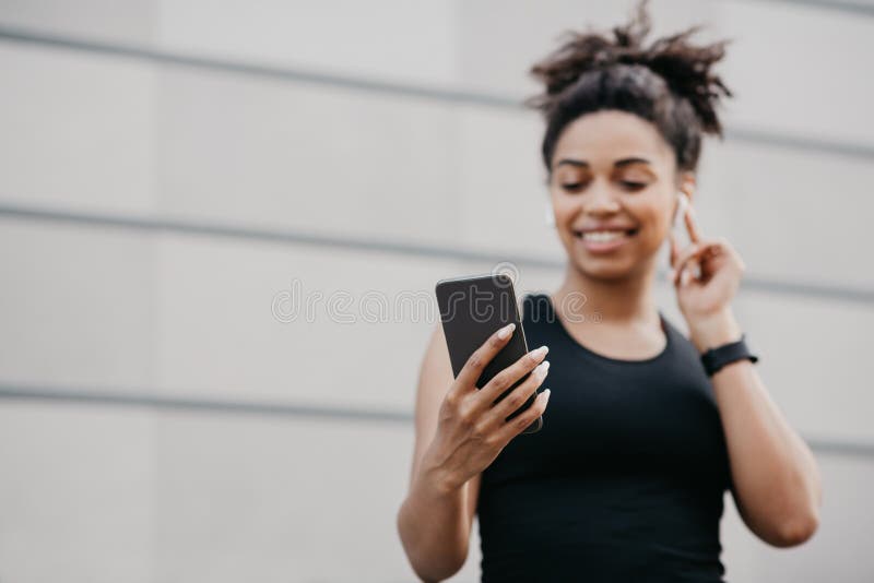 Music and running outdoors at summer. Happy african american girl in sportswear regulate wireless headphones and looks at smartphone, close up, copy space. Music and running outdoors at summer. Happy african american girl in sportswear regulate wireless headphones and looks at smartphone, close up, copy space