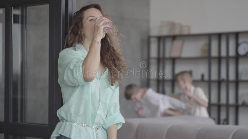 Müde junge Mutter des Porträts, die versucht, sich zu beruhigen stehend nahe der Wand mit Glas Wasser während ihre jugendlich Söh