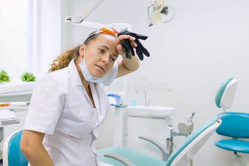 Müder Frauenzahnarzt, zahnmedizinischer Bürostuhl des Hintergrundes Weiblicher Zahnarzt, der die Handschuhe abwischen ihre Stirn lizenzfreies stockfoto Müder Frauenzahnarzt, zahnmedizinischer Bürostuhl des Hintergrundes Weiblicher Zahnarzt, der die Handschuhe abwischen ihre Stirn lizenzfreies stockfoto