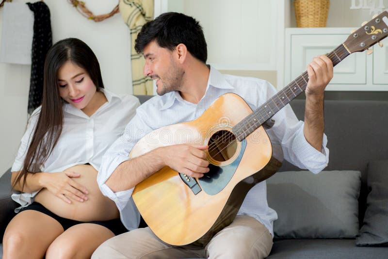 Mąż bawić się gitarę i piękną ciężarną mamy w żywym pokoju na kanapie zdjęcie stock Mąż bawić się gitarę i piękną ciężarną mamy w żywym pokoju na kanapie zdjęcie stock